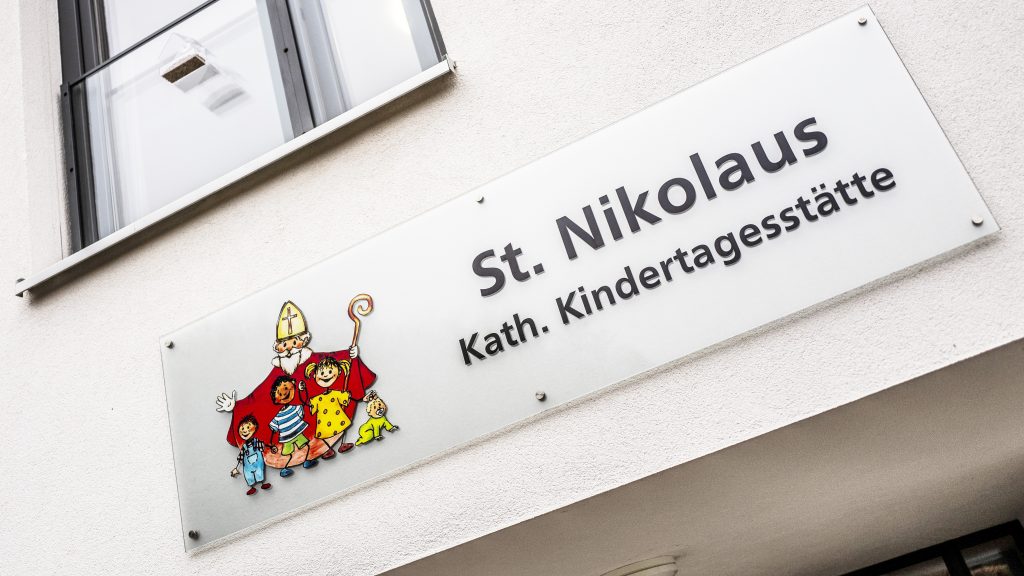 Das Schild über der Eingangstür zur Kath. Kita St. Nikolaus. Das Logo der Kita ist auf der linken Bildseite zu sehen: Der hl. Nikolaus in Mantel, Mitra und mit Hirtenstab steht hinter einigen Kindern. Daneben stehen die Worte: St. Nikolaus Kath. Kindertagesstätte.