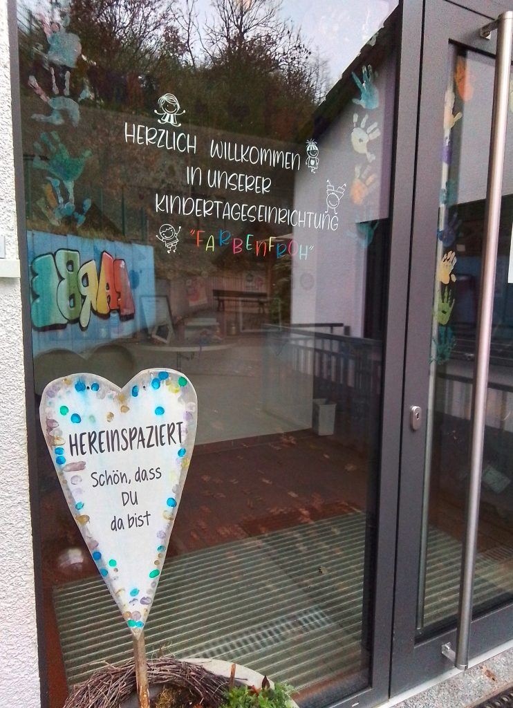 Der Eingangsbereich der Kindertagesstätte. Auf der Tür steht "Herzlich Willkommen in unserer Kindertageseinrichtung FARBENFROH". In einem Blumenkübel vor der Tür steckt ein Herz mit der Aufschrift "Hereinspaziert! Schön, dass Du da bist."