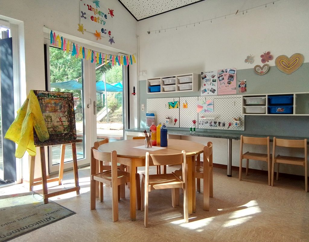 Blick in das Atelier mit Staffelei, Malkittel, einem Tisch auf dem Farben stehen und Bastelutensilien, die an einer Lochwand hängen